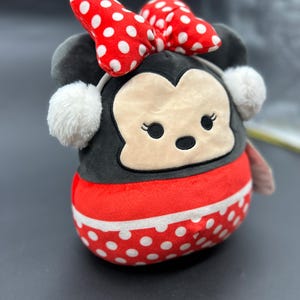 Puede incluir: Un juguete de peluche con forma de rat&oacute;n de dibujos animados con un lazo rojo y lunares blancos. El rat&oacute;n lleva una diadema blanca con un pomp&oacute;n esponjoso a cada lado. El juguete est&aacute; hecho de tela suave y de felpa y tiene una expresi&oacute;n amigable.
