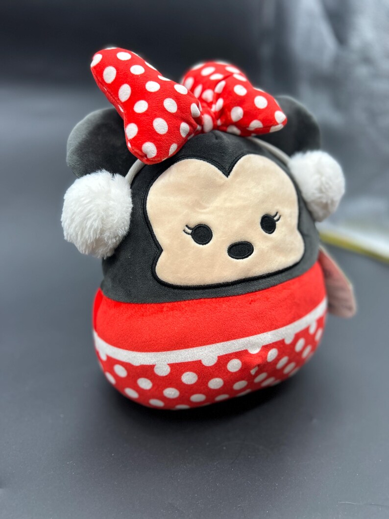Puede incluir: Un juguete de peluche con forma de rat&oacute;n de dibujos animados con un lazo rojo y lunares blancos. El rat&oacute;n lleva una diadema blanca con un pomp&oacute;n esponjoso a cada lado. El juguete est&aacute; hecho de tela suave y de felpa y tiene una expresi&oacute;n amigable.