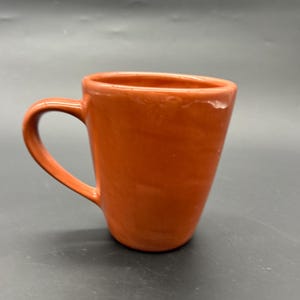 May include: A tall, orange ceramic mug with a rounded handle. The mug is smooth and has a glossy finish.