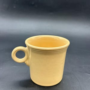Könnte beinhalten: Eine hellgelbe Keramiktasse mit einem abgerundeten Henkel. Die Tasse hat eine zylindrische Form mit einem leicht ausgestellten Rand. Die Tasse ist aus Keramik gefertigt und hat eine glatte, glänzende Oberfläche. Die Tasse ist für Heißgetränke konzipiert.