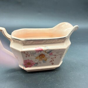 May include: A vintage, pale pink gravy boat with a floral pattern. The boat has a rectangular shape with a handle and a spout. The floral design includes pink, yellow, and blue flowers. The rim is trimmed with a silver-coloured line.