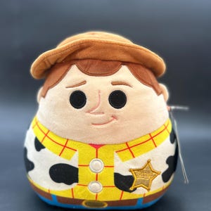 May include: A plush toy of Woody from Toy Story. He is wearing his signature cowboy hat, vest, and sheriff's badge. He has a brown hat, a yellow shirt with red squares, a black and white cow pattern, and a blue belt.