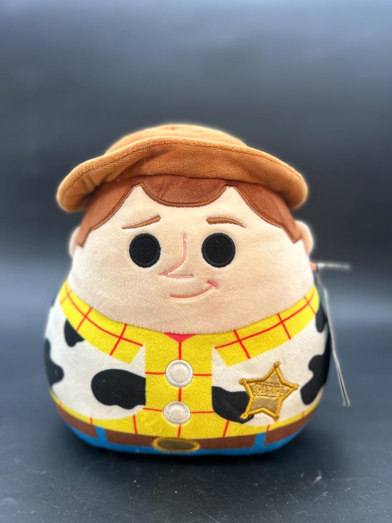 May include: A plush toy of Woody from Toy Story. He is wearing his signature cowboy hat, vest, and sheriff's badge. He has a brown hat, a yellow shirt with red squares, a black and white cow pattern, and a blue belt.