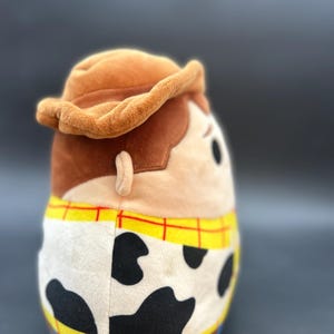 May include: A brown plush toy shaped like Woody from Toy Story. The toy is wearing a brown cowboy hat, a yellow and red checked shirt, and a brown belt with a silver buckle. The toy has a white and black cow print pattern on the bottom half of its body.