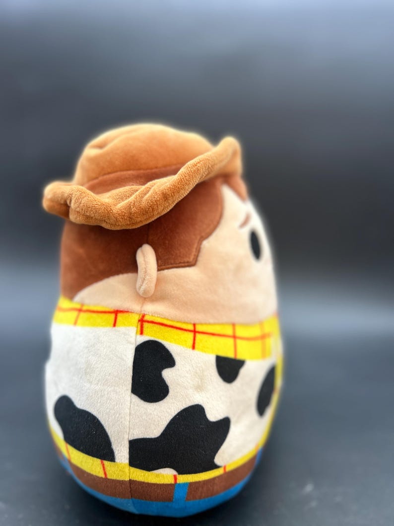 May include: A brown plush toy shaped like Woody from Toy Story. The toy is wearing a brown cowboy hat, a yellow and red checked shirt, and a brown belt with a silver buckle. The toy has a white and black cow print pattern on the bottom half of its body.