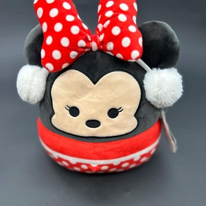 Puede incluir: Un juguete de peluche con forma de rat&oacute;n de dibujos animados con un lazo rojo y lunares blancos. El rat&oacute;n lleva una bufanda de rayas rojas y blancas y tiene orejeras de piel blanca.
