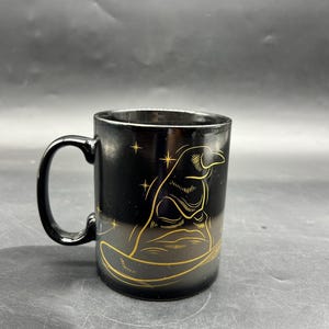May include: Black ceramic mug with a gold illustration of a wizard's hat and stars. The mug has a black handle and a gold rim. The design is on the front of the mug.