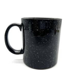 May include: Black speckled ceramic mug with a large handle. The mug has a speckled pattern of small white dots on a black background. The mug is cylindrical and sits on a white surface.