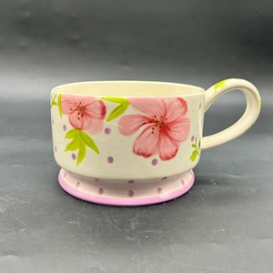 May include: Cream-colored ceramic mug with a floral design. The mug features pink flowers, green leaves, and purple polka dots. The base is light pink with a darker pink trim, and it has a curved handle.