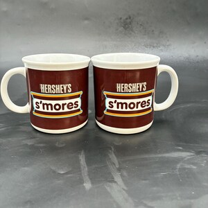 Może przedstawiać: Dwa kubki Hershey's s'mores. Kubki są ciemnobrązowe z białymi wnętrzami i uchwytami. Logo Hershey's i tekst s'mores są w białym i pomarańczowym wzorze. Kubki są wykonane z ceramiki.