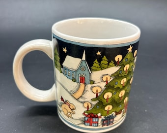 Pode incluir: Uma caneca de cerâmica com alça e borda brancas. A caneca apresenta uma cena de inverno com uma igreja, árvore de Natal, presentes e um coelho. O design é contra um fundo azul escuro com estrelas.