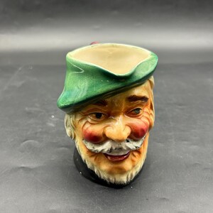 Puede incluir: Una taza de cerámica con forma de cabeza de hombre, con un sombrero verde, bigote blanco y mejillas rojas. La taza tiene un cuello negro y un interior beige. El diseño es detallado y caprichoso, lo que sugiere un artículo de colección o decorativo.