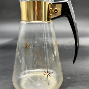 May include: A clear glass pitcher with a gold band around the top and a black plastic handle. The pitcher has a starburst design on the side.