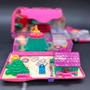 以下が含まれることがあります： ピンクと緑色のヴィンテージミニチュアプレイセット。詳細な内部構造が明らかになっています。紫色の屋根の小さな家、緑の丘、ケンケンパのコートが含まれています。プレイセットは折りたたんで閉じられるように設計されています。