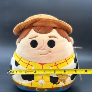 May include: A plush toy of Woody from Toy Story. He is wearing his signature cowboy hat, vest, and sheriff's badge. The toy is approximately 18 cm tall.
