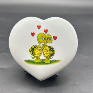 May include: A heart-shaped ceramic trinket box with a white glossy finish. The lid features a cartoon illustration of two yellow turtles embracing, with red hearts floating above them. The turtles have green and yellow shells and are standing on green grass.