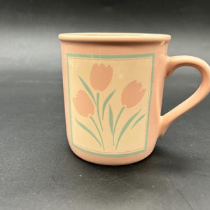 May include: A pink ceramic mug with a curved handle. The mug features a rectangular design with three pink tulip flowers and green stems on a cream background, framed by a light green border. The mug is set against a dark background.