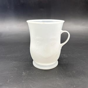May include: A translucent white plastic mug with a handle and a smiling face design. The mug has a flared top and a circular base. The face is subtly embossed on the front. The mug is on a dark surface.
