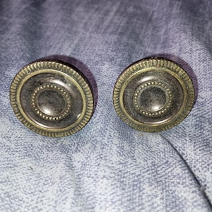 May include: Two antique-style cabinet knobs. Each knob is round with a dark center and a decorative, ridged outer edge. The knobs have a bronze-like finish and are set against a textured, blue-gray background.