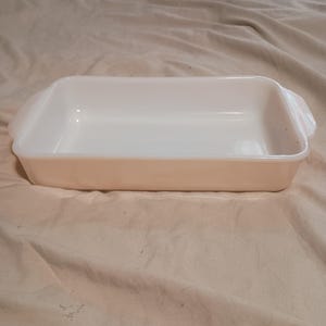 May include: A rectangular, off-white baking dish with rounded corners and handles. The dish is empty and appears to be made of ceramic or glass. The background is a neutral-toned fabric.