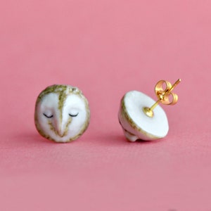 Pode incluir: Um par de brincos de cerâmica branca em forma de coruja com postes dourados. As corujas têm os olhos fechados e detalhes castanhos.