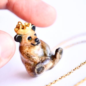 May include: A ceramic bear pendant with a gold crown, wearing a gold chain necklace. The bear is brown and black with a light brown belly and is sitting up with its paws tucked in.