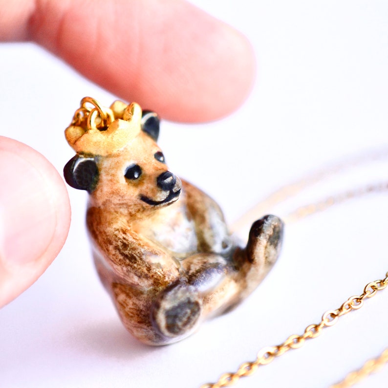 May include: A ceramic bear pendant with a gold crown, wearing a gold chain necklace. The bear is brown and black with a light brown belly and is sitting up with its paws tucked in.