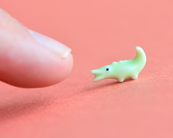 Könnte beinhalten: Eine kleine grüne Keramik-Alligator-Figur mit schwarzen Augen auf einem rosafarbenen Hintergrund. Ein Finger zeigt auf den Alligator.