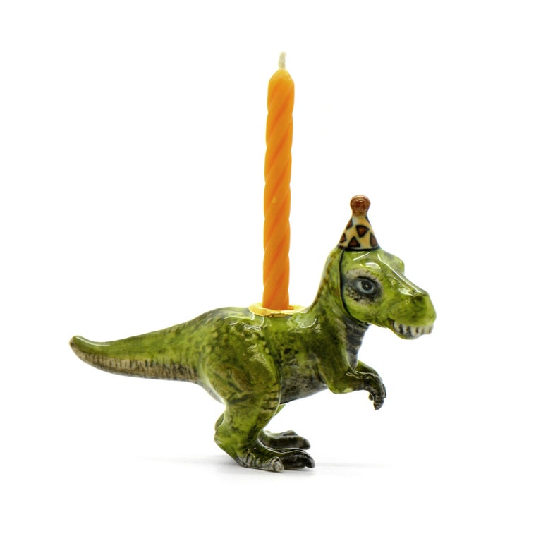 Pode incluir: Um porta-velas de anivers&aacute;rio de cer&acirc;mica verde Tyrannosaurus Rex com um chap&eacute;u de festa e uma vela laranja. O dinossauro tem um acabamento texturizado detalhado. A vela tem cerca de 15 cm de altura.