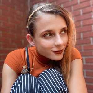 May include: A person wearing a brown turtleneck shirt with a gold chain necklace featuring a small animal pendant. They are also wearing black and white striped pants and a gold headband.