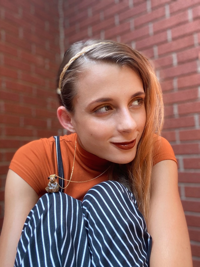 May include: A person wearing a brown turtleneck shirt with a gold chain necklace featuring a small animal pendant. They are also wearing black and white striped pants and a gold headband.