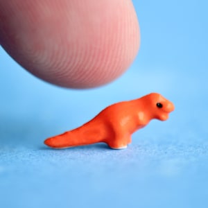 May include: A small, orange dinosaur figurine with black eyes and white feet. The miniature sculpture is set against a light blue background. The figurine is positioned near a fingertip.