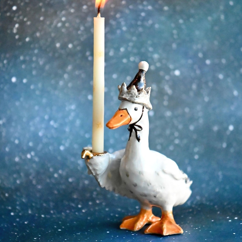 May include: A decorative white goose ornament, wearing a crown and holding a lit candle. The goose has orange feet and beak, with a black bow tie. The candle is ivory, and the background is a speckled blue.