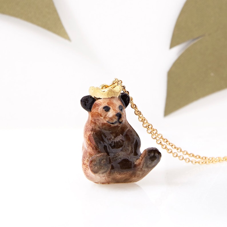 May include: A brown ceramic bear pendant with a gold crown on its head, hanging from a gold chain necklace.