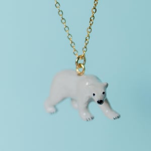 May include: A gold chain necklace featuring a white polar bear pendant. The polar bear has black eyes and paws. The background is a light blue colour.