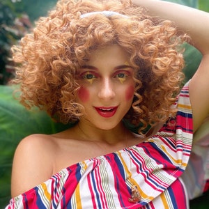 May include: A woman with curly red hair wearing a red, white, and blue striped off-the-shoulder top with a gold necklace and a small pendant. She has bright red blush and gold glitter on her eyelids.