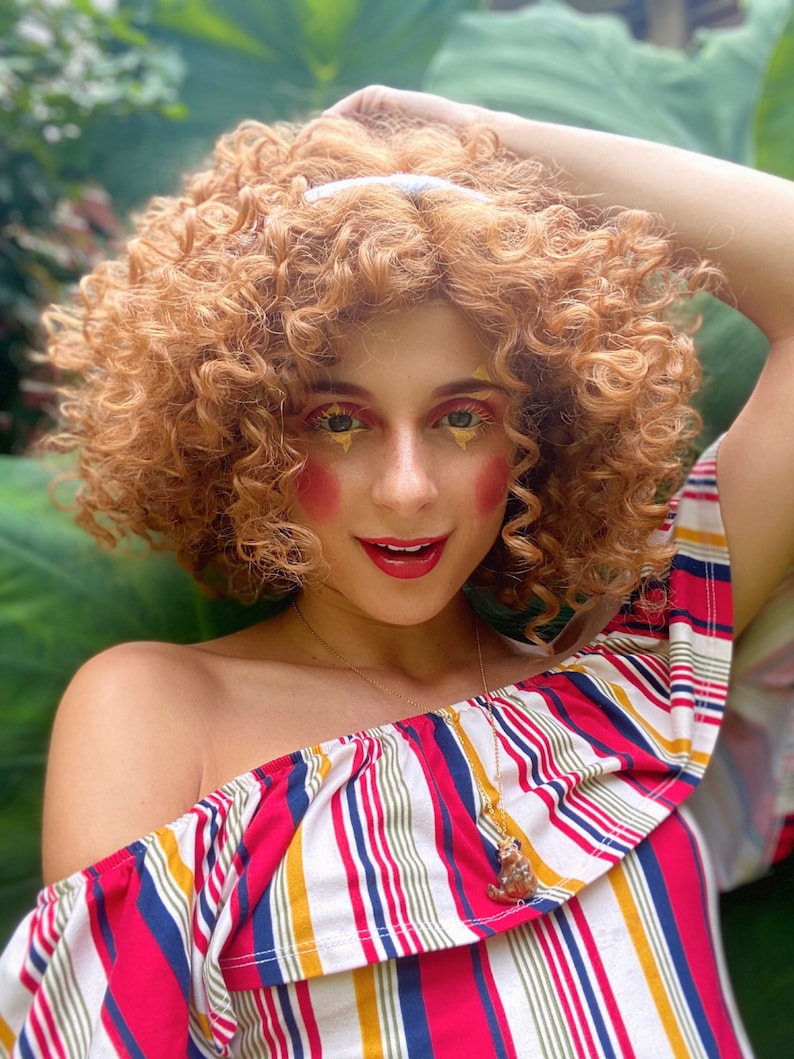 May include: A woman with curly red hair wearing a red, white, and blue striped off-the-shoulder top with a gold necklace and a small pendant. She has bright red blush and gold glitter on her eyelids.