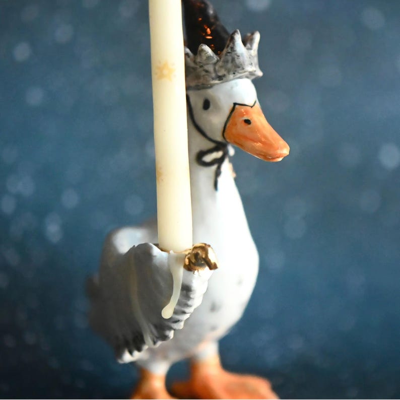 May include: A decorative ceramic goose candle holder. The goose is white with grey wings, an orange beak and feet, and a silver crown. It holds a cream-coloured candle. The background is a blurred blue, with a black bow tie.