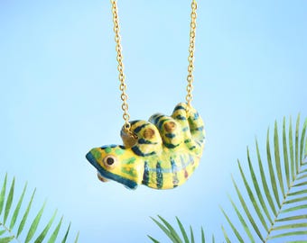 May include: A gold chain necklace with a yellow, blue and green ceramic chameleon pendant. The chameleon is hanging upside down.