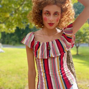 May include: A woman with curly brown hair wears a red, white, and blue striped off-the-shoulder top with a ruffled neckline. She is wearing a gold necklace with a pendant.