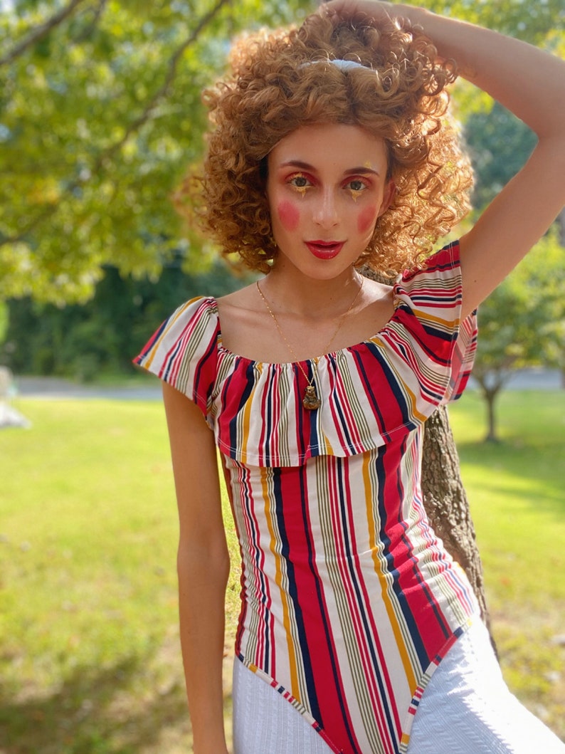 May include: A woman with curly brown hair wears a red, white, and blue striped off-the-shoulder top with a ruffled neckline. She is wearing a gold necklace with a pendant.