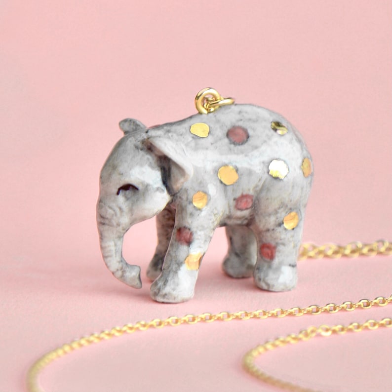 May include: A gold chain necklace with a ceramic elephant pendant. The elephant is gray with gold and red polka dots.