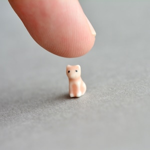 May include: A small, light pink ceramic figurine of a cat sitting on a gray surface. The cat has black eyes and a white chest.