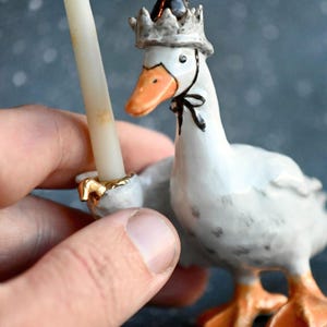 May include: A white ceramic goose figurine wearing a crown and a black bow tie, holding a candle. The goose has an orange beak and feet, with gold accents on the candle holder. A whimsical decorative item.