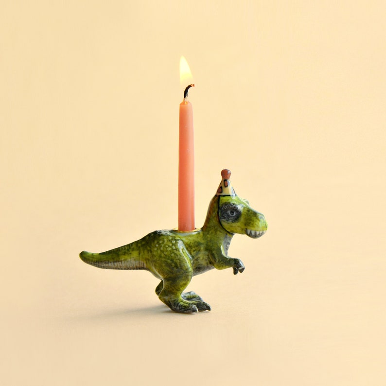 Pode incluir: Um casti&ccedil;al em forma de dinossauro verde com um chap&eacute;u de festa, segurando uma vela rosa acesa. O casti&ccedil;al est&aacute; em um fundo amarelo claro. O dinossauro tem uma apar&ecirc;ncia detalhada e texturizada.
