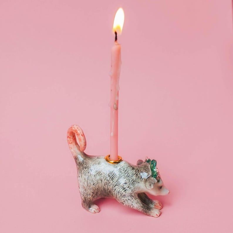 Op de afbeelding: Een keramische kandelaar in de vorm van een opossum, met een brandende roze kaars. De opossum is grijs en wit met een gekrulde staart en een kleine groene bloemenkrans. De kaars staat in een goudkleurige ring.