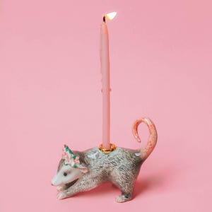 Op de afbeelding: Een keramische kandelaar in de vorm van een opossum met een brandende roze kaars. De opossum is grijs en wit met een bloemenkrans. De kaars heeft druipend was. De houder heeft een gouden ring.
