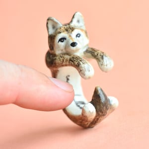 May include: A ceramic wolf ring with brown and white fur details. The wolf is curled up with its tail wrapped around its body. The ring is designed to be worn on the finger.