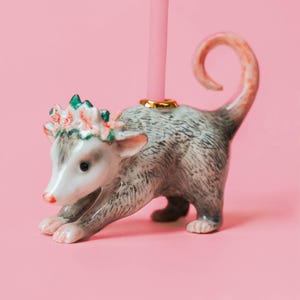 Op de afbeelding: Een keramische opossum kaarsenhouder met een roze kaars. De opossum is grijs en wit met een roze staart en oren, en een bloemenkrans. De kaarsenhouder heeft een goudkleurige basis. De achtergrond is roze.