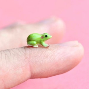 May include: A small green ceramic frog figurine with pink spots on its face. The frog is sitting on a finger.